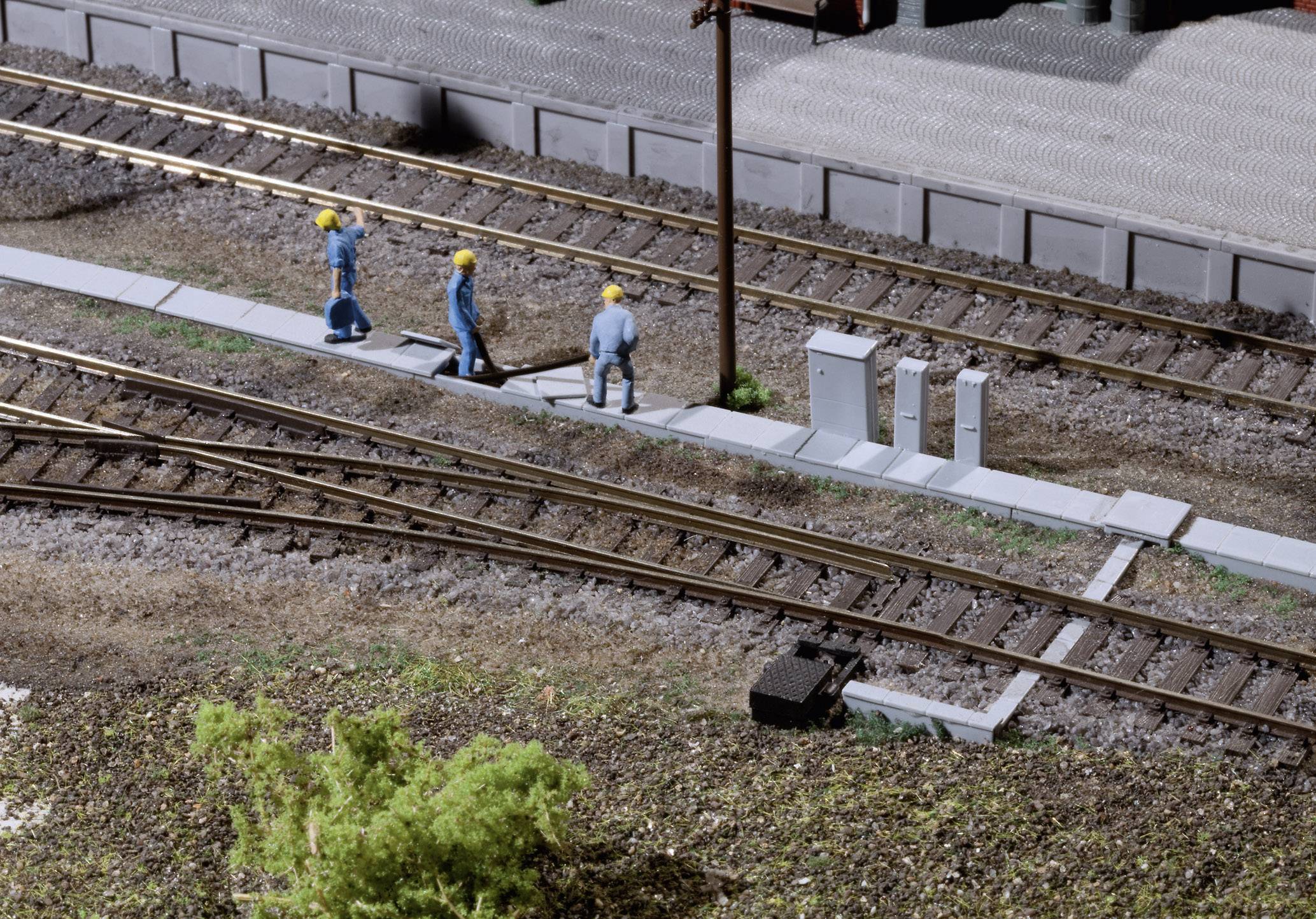 Tri miniatúrne figúrky s farebnými žltými helmami pracujú vedľa železničných koľají. Scéna zobrazuje realistické prostredie železničných robotníkov.