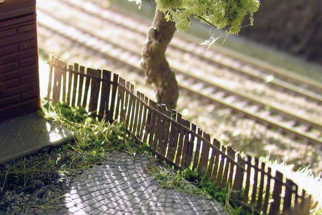 Modelová krajina so šikmým dreveným plotom, stromom a železničnými koľajami v pozadí; zobrazuje idylickú scénu v miniaturizovanom formáte.