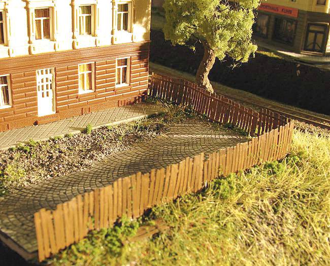 Model železnice so starobylou tehlovou budovou, starým stromom a dreveným plotom na trávnatom teréne.