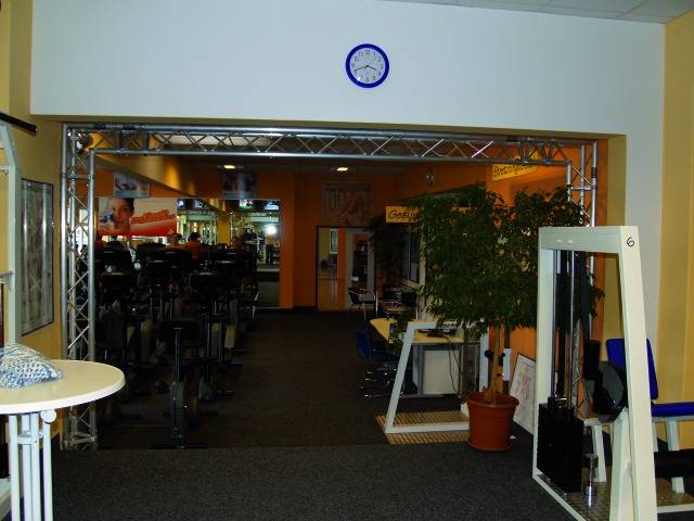 Interiér fitnescentra s viacerými fitness zariadeniami, žltými stenami, zelenou rastlinou a nástenými hodinami nad vstupom.