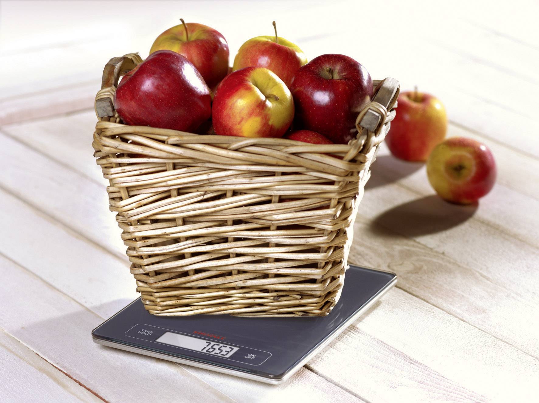 Košík s červenými jablkami stojí na digitálnej kuchynskej váhe, ktorá ukazuje hmotnosť 765 gramov.