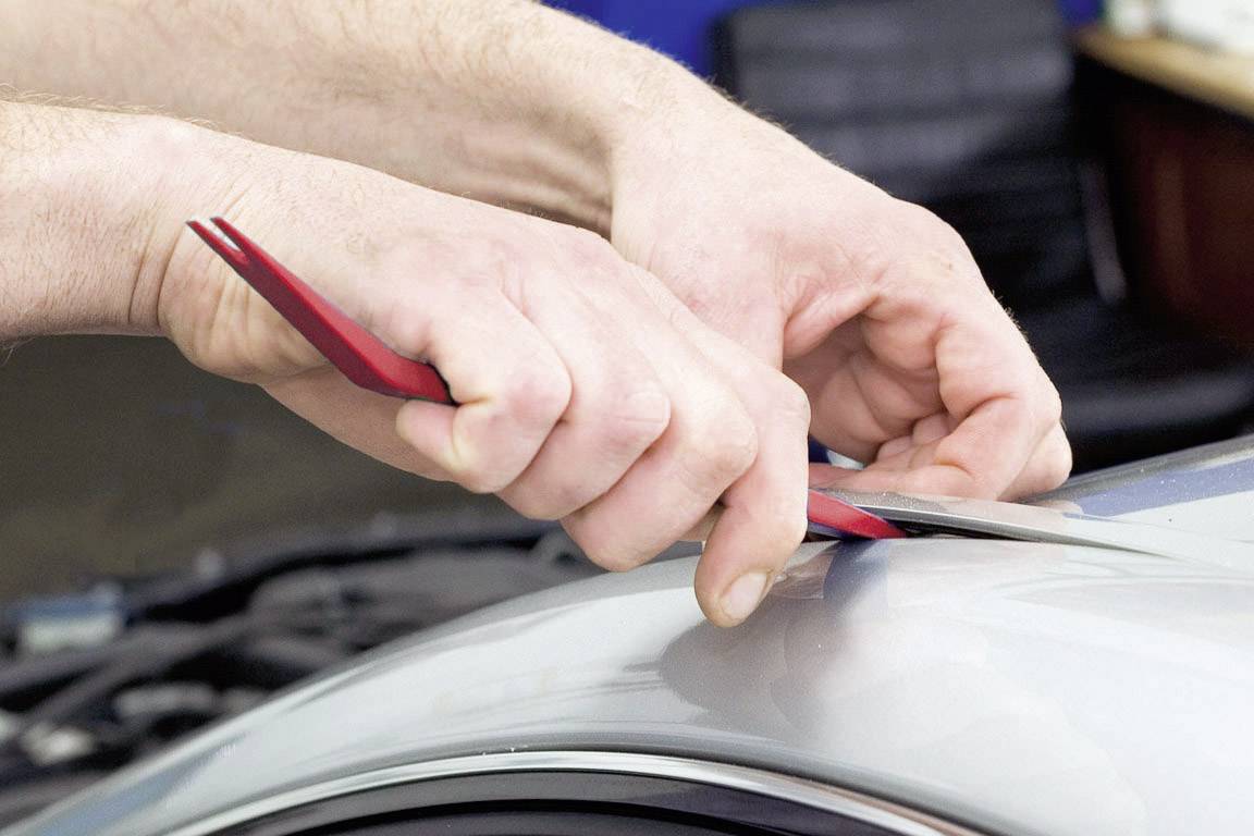 Osoba odstraňuje pomocou červeného nástroja preliačinu zo strieborného automobilu. Detailný záber zachytáva precíznu prácu na vozidle.