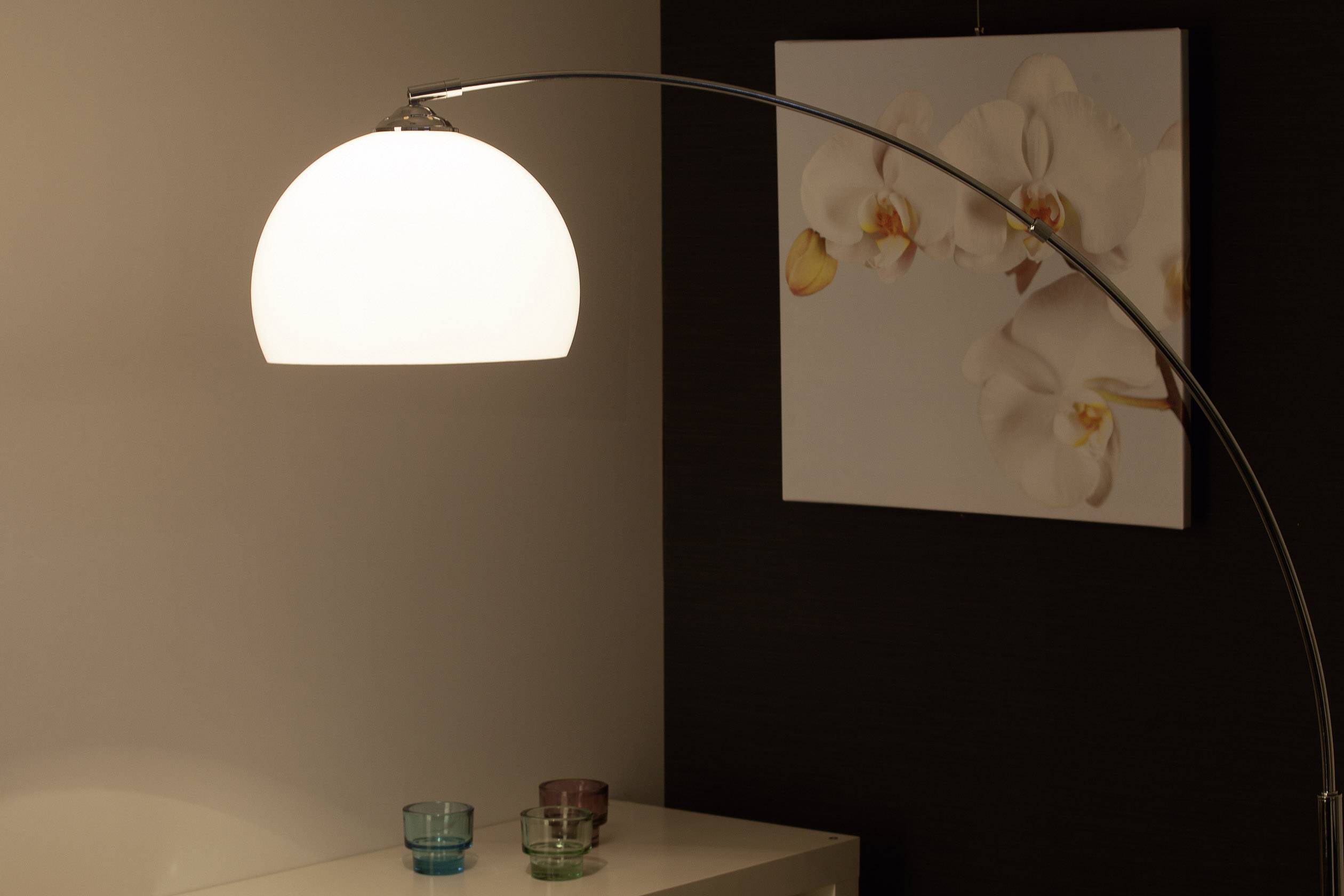 Moderná stojanová lampa s ohnutým ramenom a bielym tienidlom stojí pred tmavým pozadím s obrazom orchidey.