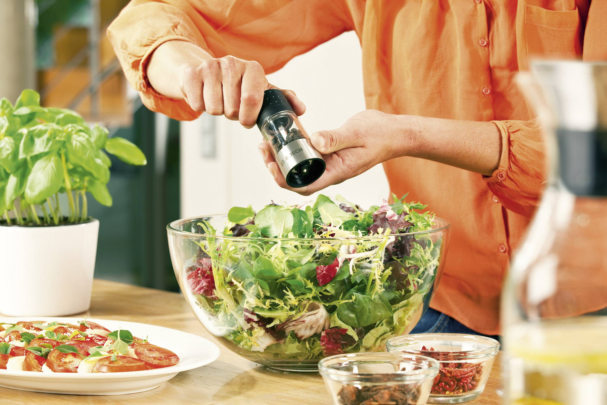 Osoba v oranžovej košeli korení čerstvý listový šalát v sklenenej miske čiernym korením. Vedľa stojí hrnček čerstvej bazalky.