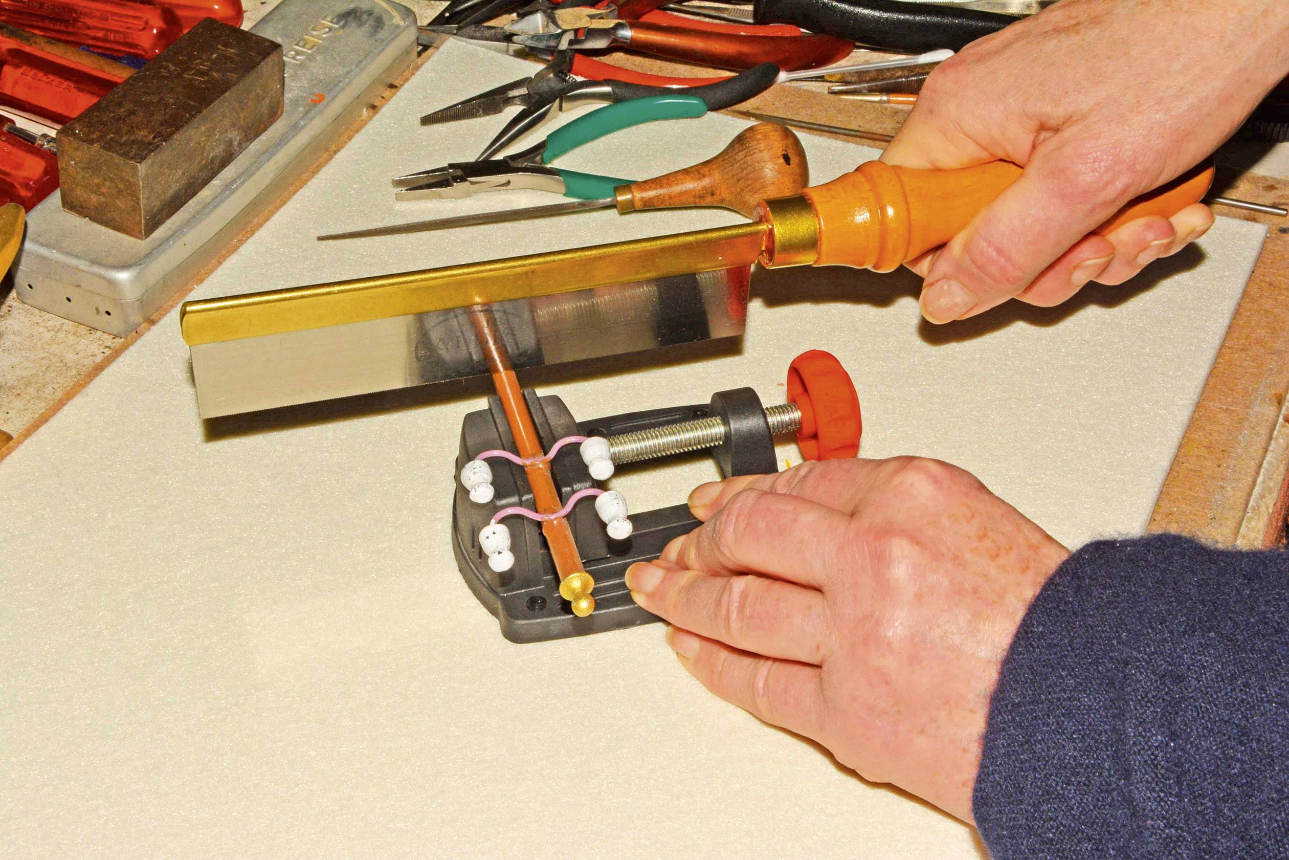 Osoba píli tenký medený drôt na malej pracovnej lavici pomocou kovovej píly. V pozadí ležia nástroje ako kliešte a skrutkovače.