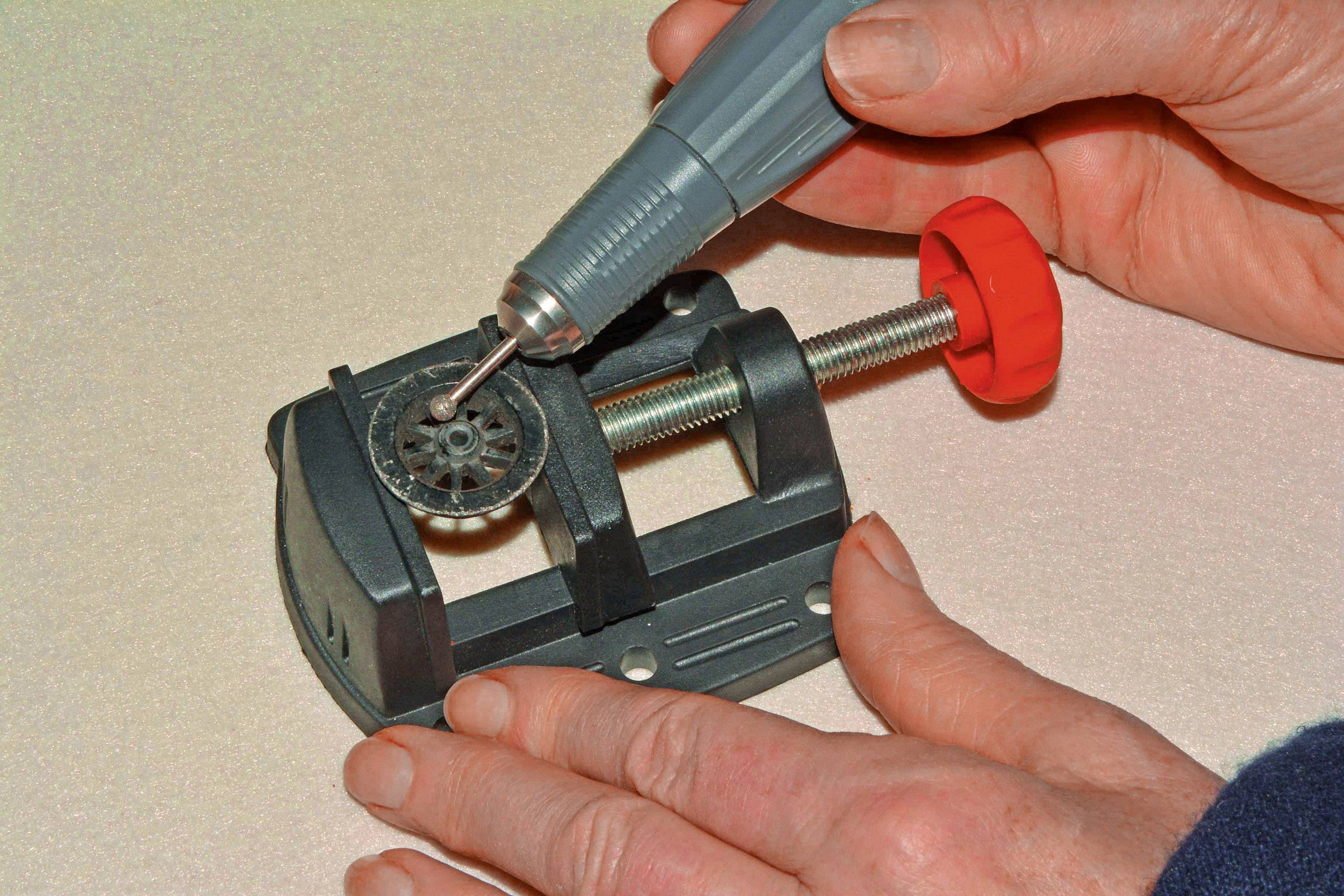 Jedna ruka drží rotujúci nástroj a pracuje na kovovej súčiastke upevnenej vo zveráku, pričom druhá ruka pridržiava zverák v stabilnej polohe.