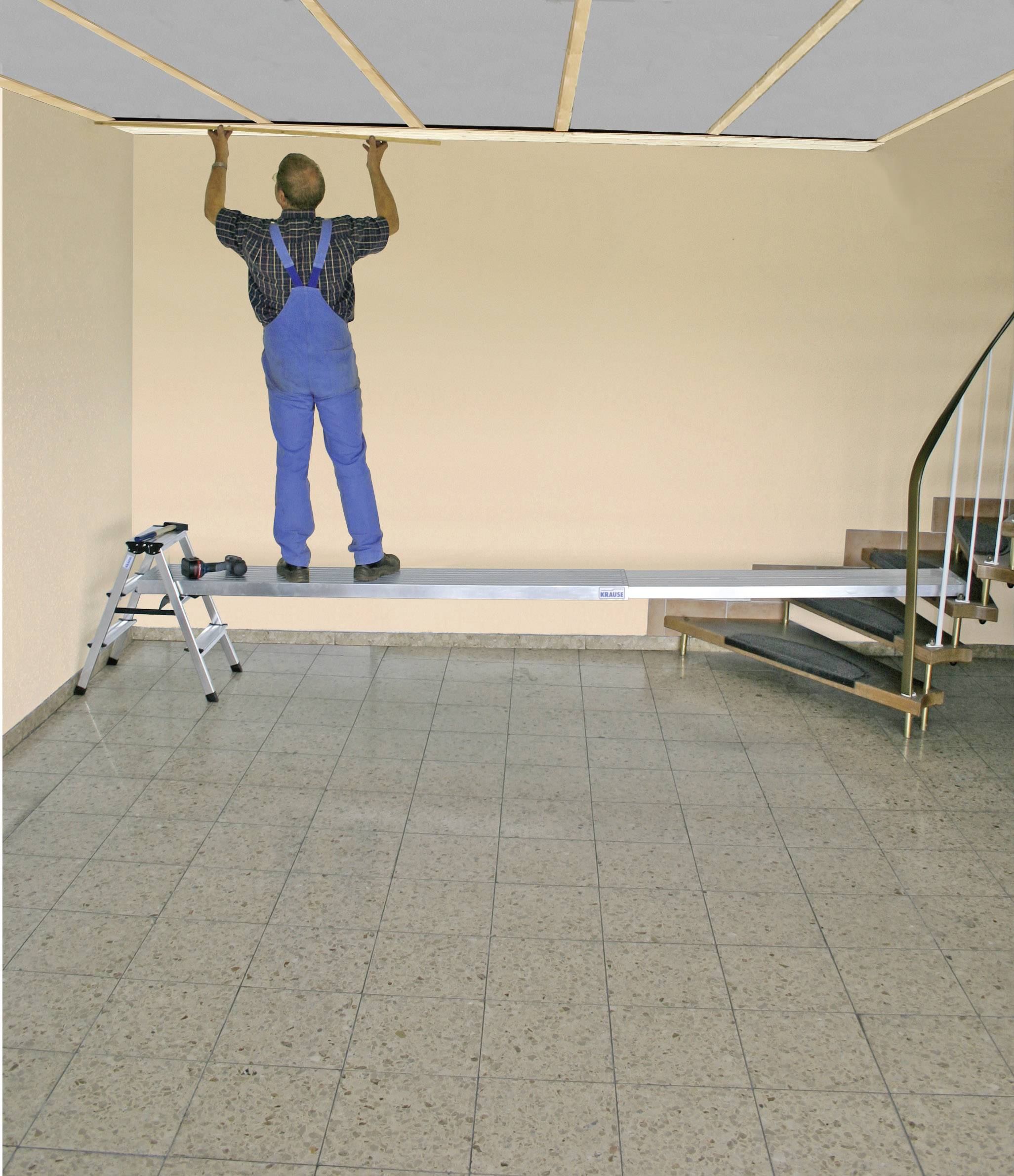 Remeselník v modrom montérkovom overale inštaluje stropný obklad v miestnosti so schodiskom do špirály a dlaždicovou podlahou.