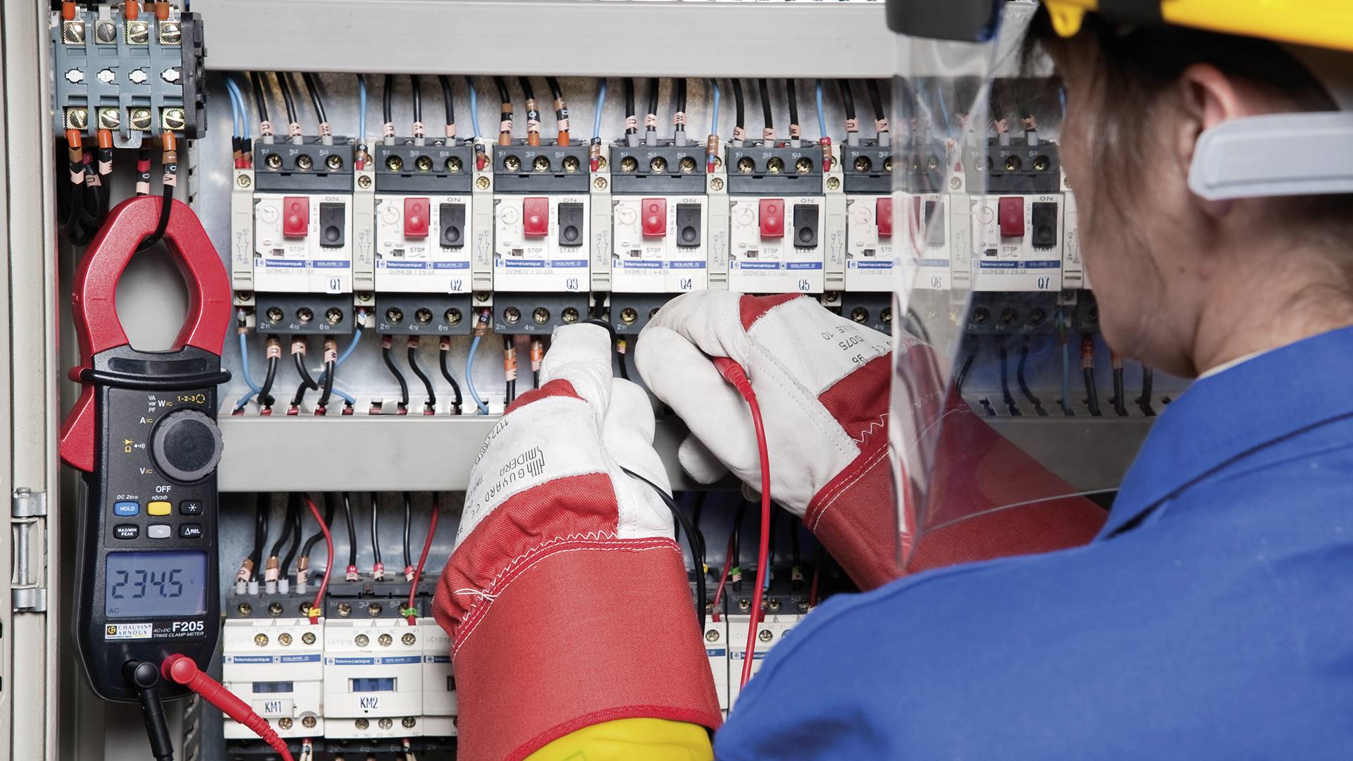 Oseba v zaščitni opremi dela na električni omari in meri električne vrednosti z multimetrom, pri čemer je prikazana vrednost 234,6.