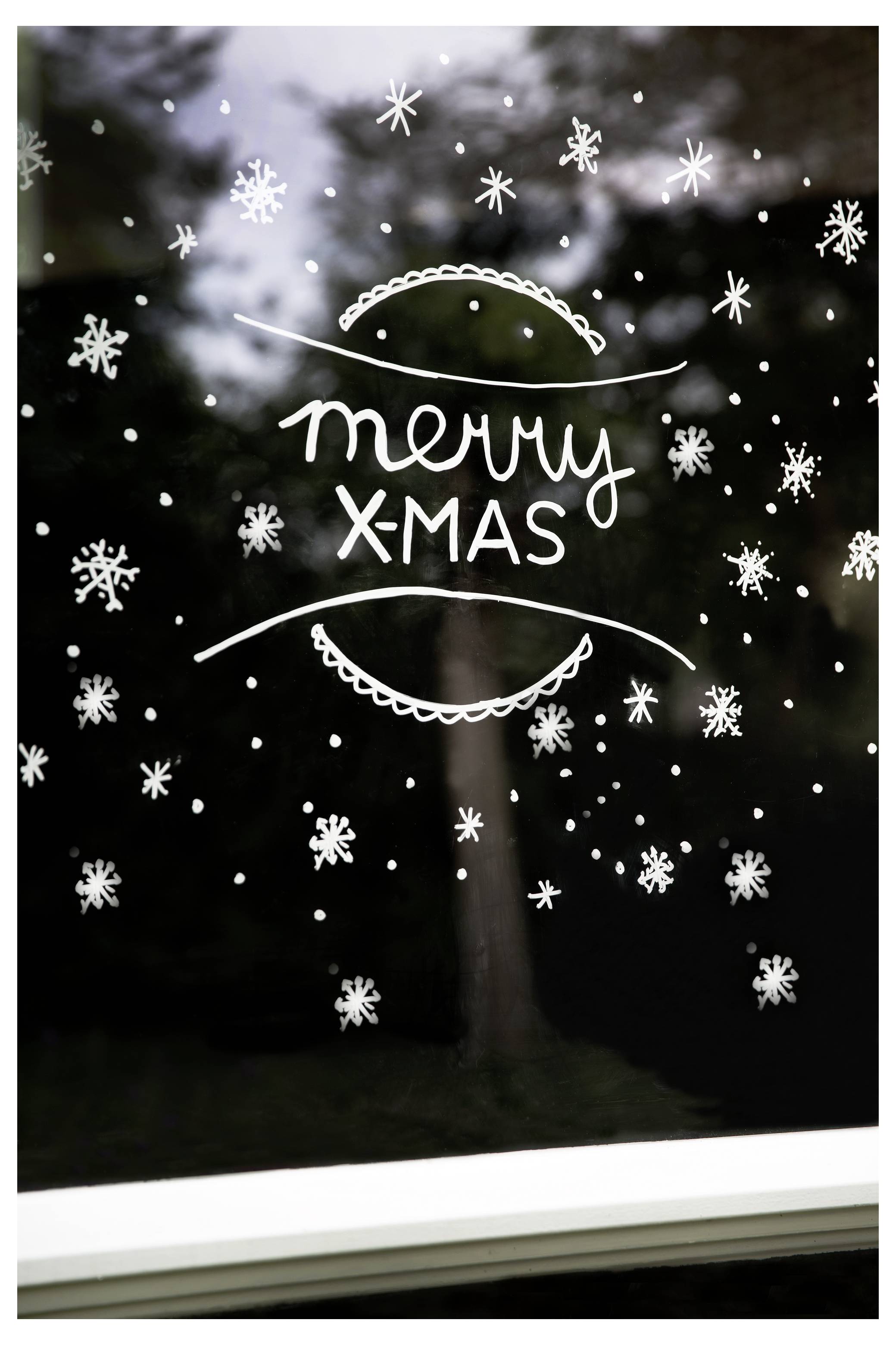 Božična okrasitev okna z ročno narisanimi snežinkami in besedami 'veseli X-MAS' na temnem ozadju.