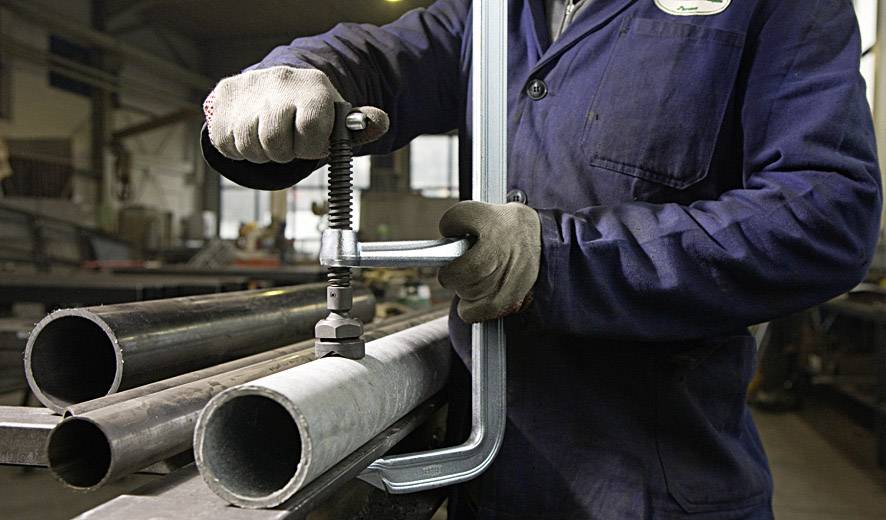 Oseba v delovni obleki uporablja C-objemko za pritrjevanje kovinskih cevi v delavnici. V ozadju so vidna še druga industrijska orodja.