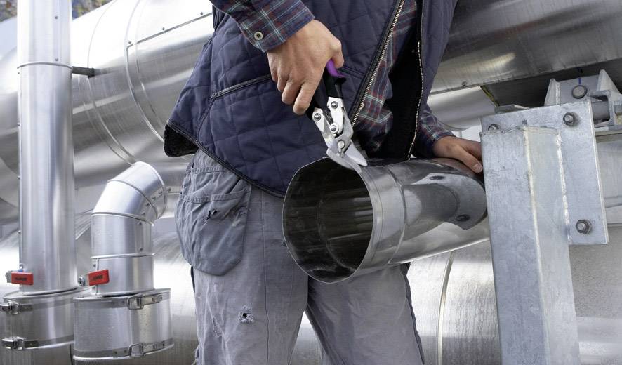 Oseba s kovinskimi škarjami reže okroglo kovinsko cev poleg industrijskih objektov. Oseba nosi delovno obleko.