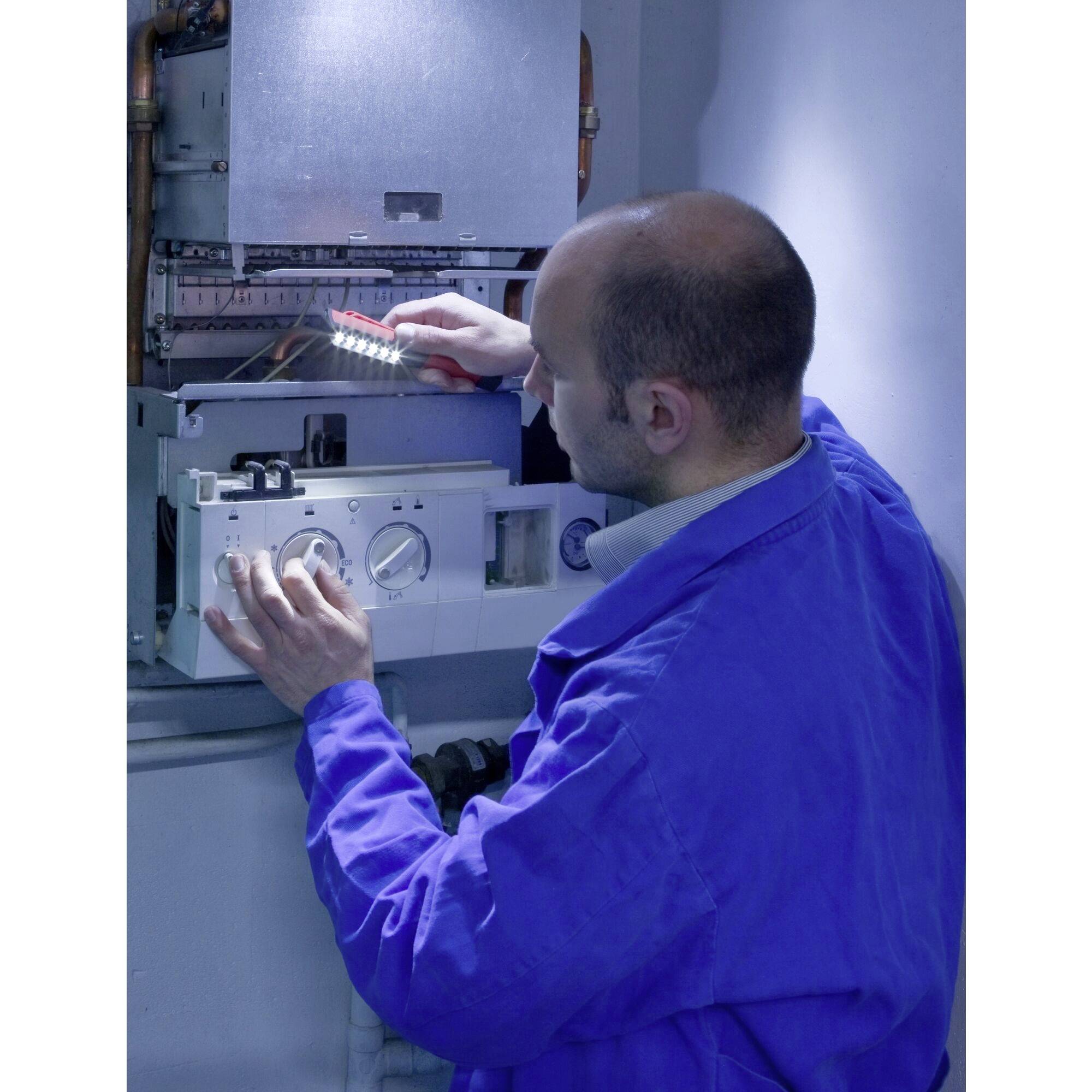 Oseba popravlja grelni kotel, oblečena je v moder delovni kombinezon in drži žepno svetilko, da razsvetljuje delovno mesto.