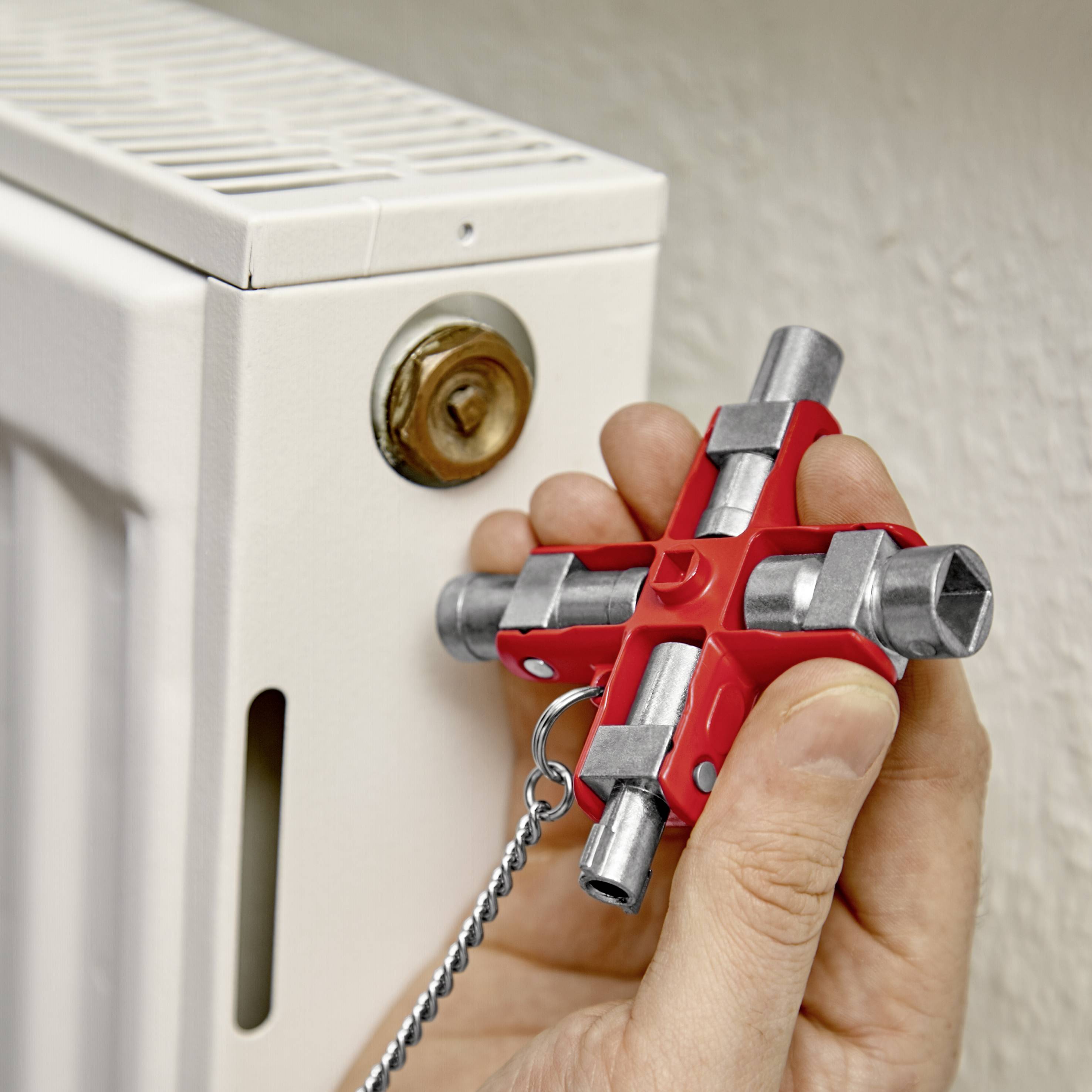 Oseba drži ključ za radiator in odpira ventil za izpust zraka na radiatorju. Ključ ima štiri različne konce.