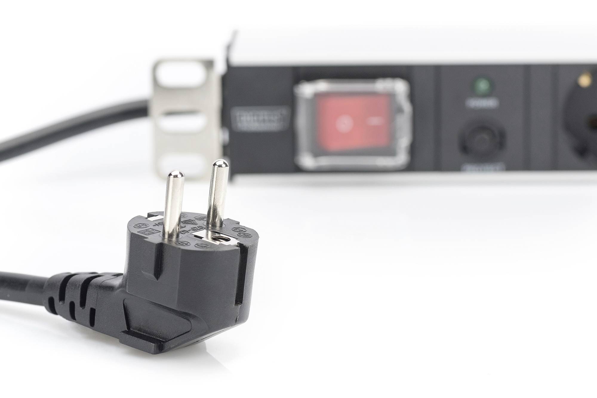 Črn električni vtič v ospredju, nejasno večtaktno razdelilno letev s stikalom v ozadju. Simbolizira elektriko in možnosti priključevanja.
