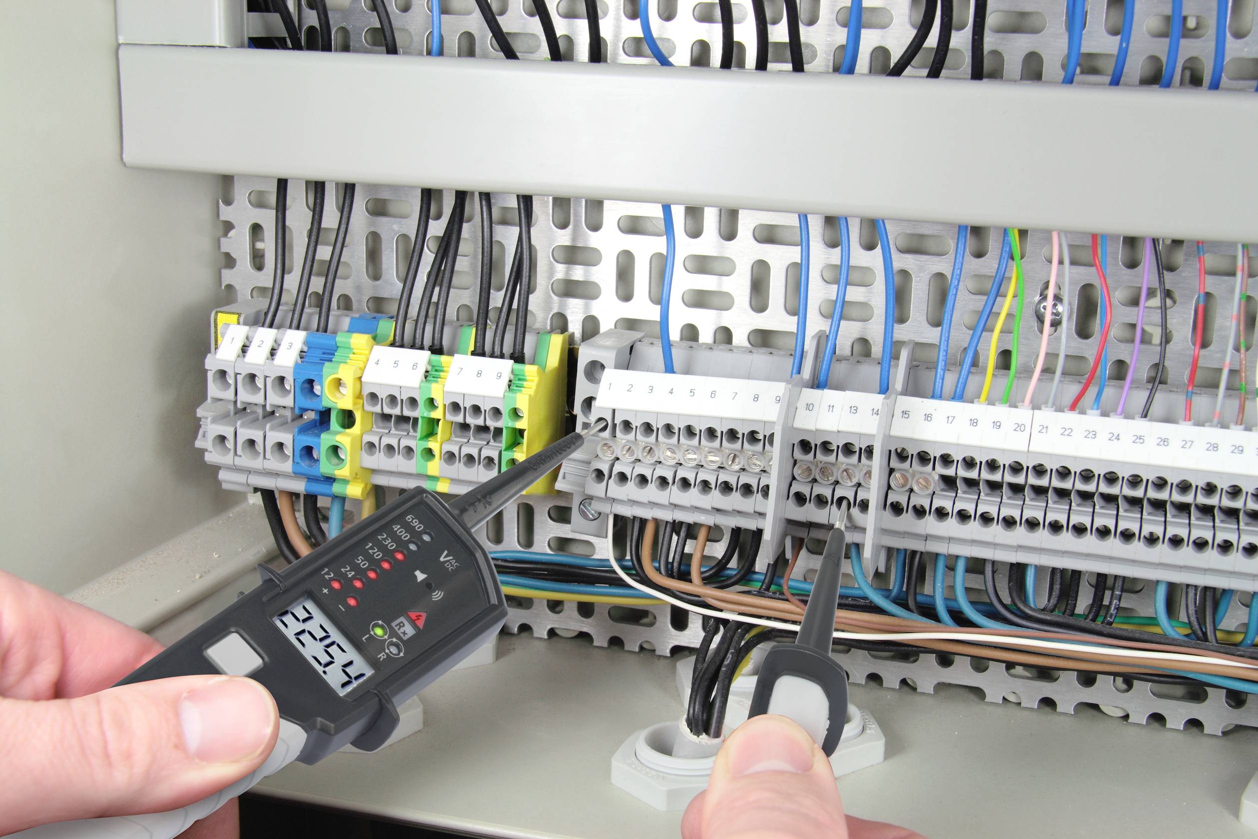 Oseba meri napetost na elektrorazdelilniku, merilna naprava prikazuje '225,2 V'. Kabli in priključne sponke so vidni.