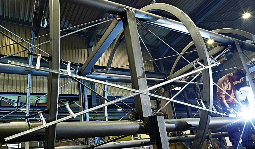 Oseba vara v veliki industrijski napravi s jeklenim okvirjem in modrimi konstrukcijskimi elementi v delavnici.