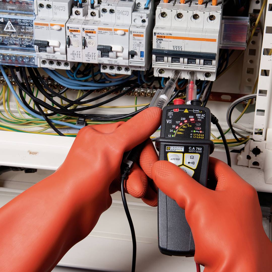 Električar z oranžnimi rokavicami meri napetost v elektrodozornem omarici. Prikazuje natančno delo z merilno napravo.