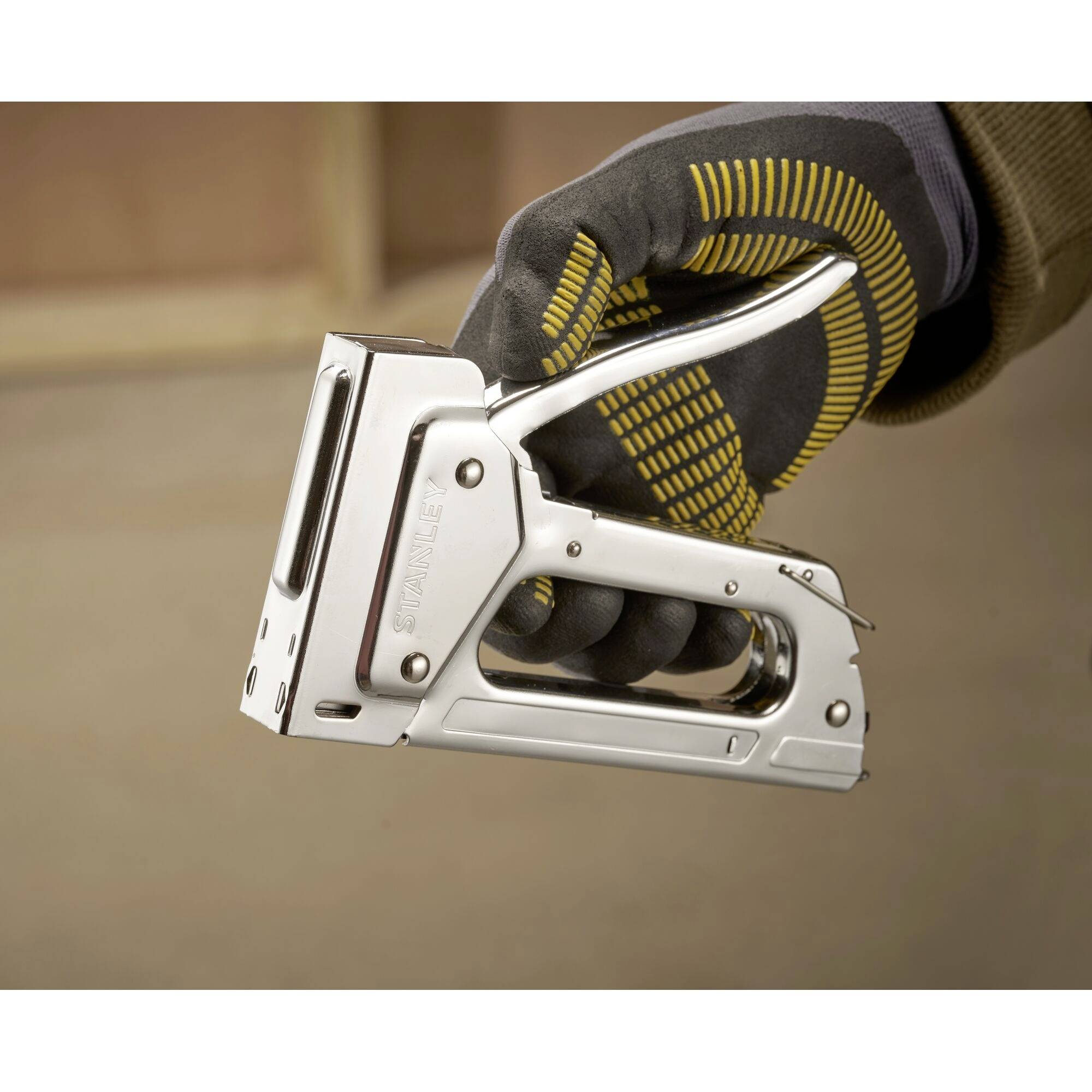 Oseba drži srebrn spenjalnik v roki, ki je v črni rokavici z rumenimi črtami. Ozadje je zamegljeno.