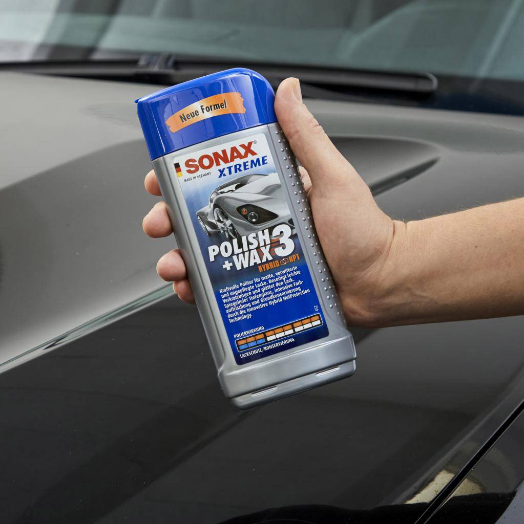 Ena roka drži steklenico 'Sonax Xtreme Polish + Wax 3', avtopolirnega in voskanega izdelka. Ozadje prikazuje temno vozilo.
