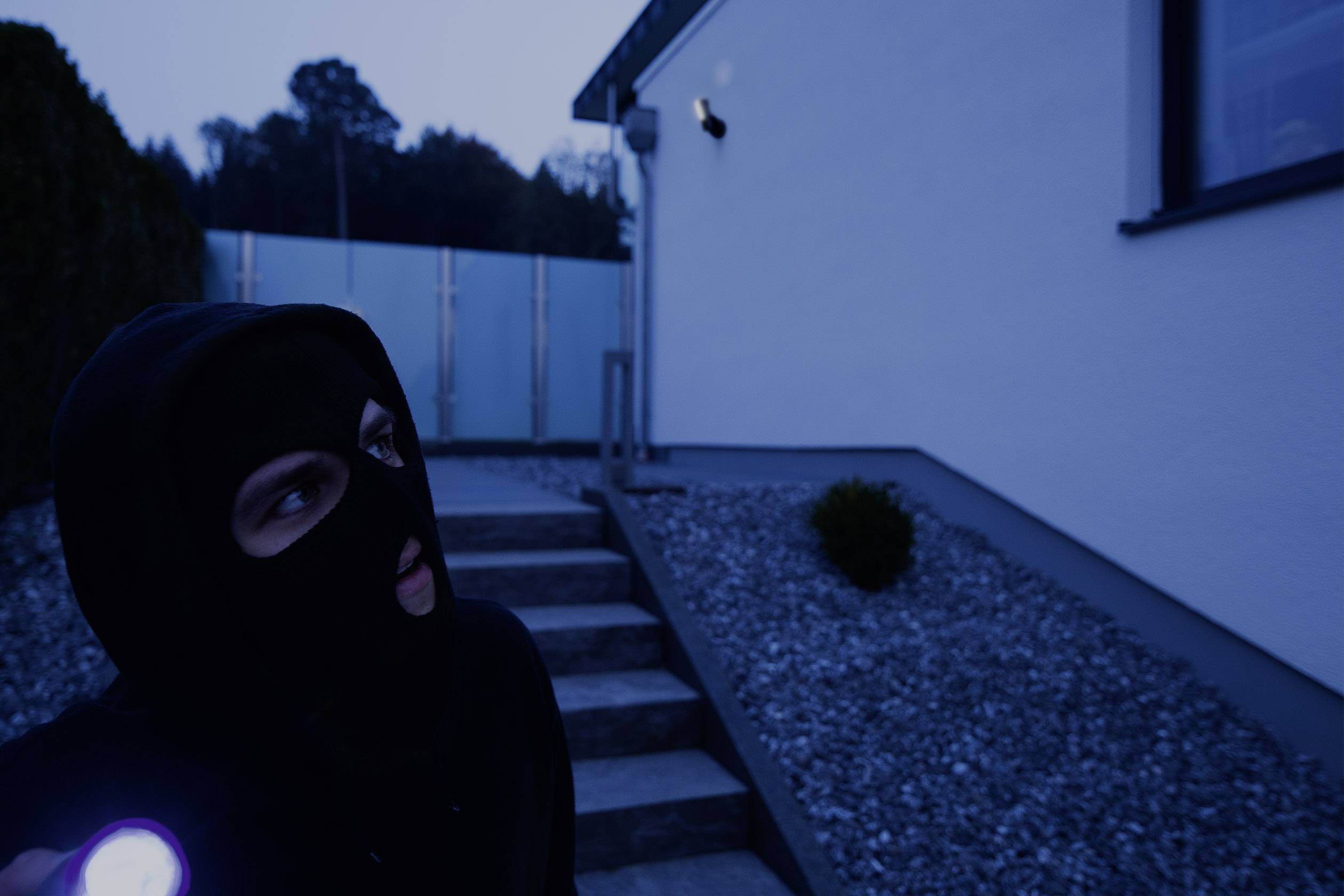 Mask­irana oseba stoji ponoči pred hišo in sveti z žepno svetilko.