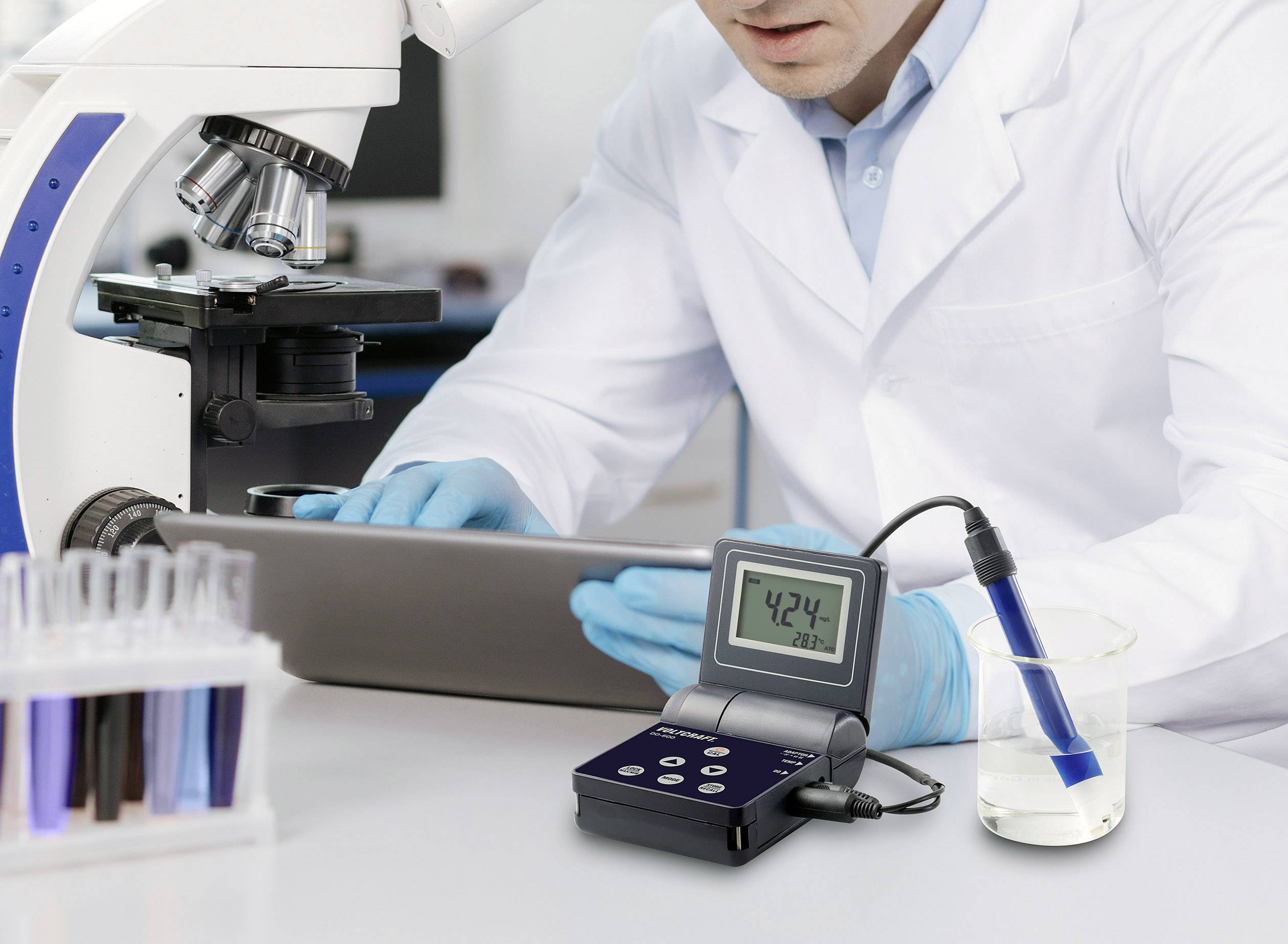 Znanstvenik v laboratoriju dela z mikroskopom, ob njem digitalna merilna naprava, ki prikazuje 42,4, in elektroda v čaši.