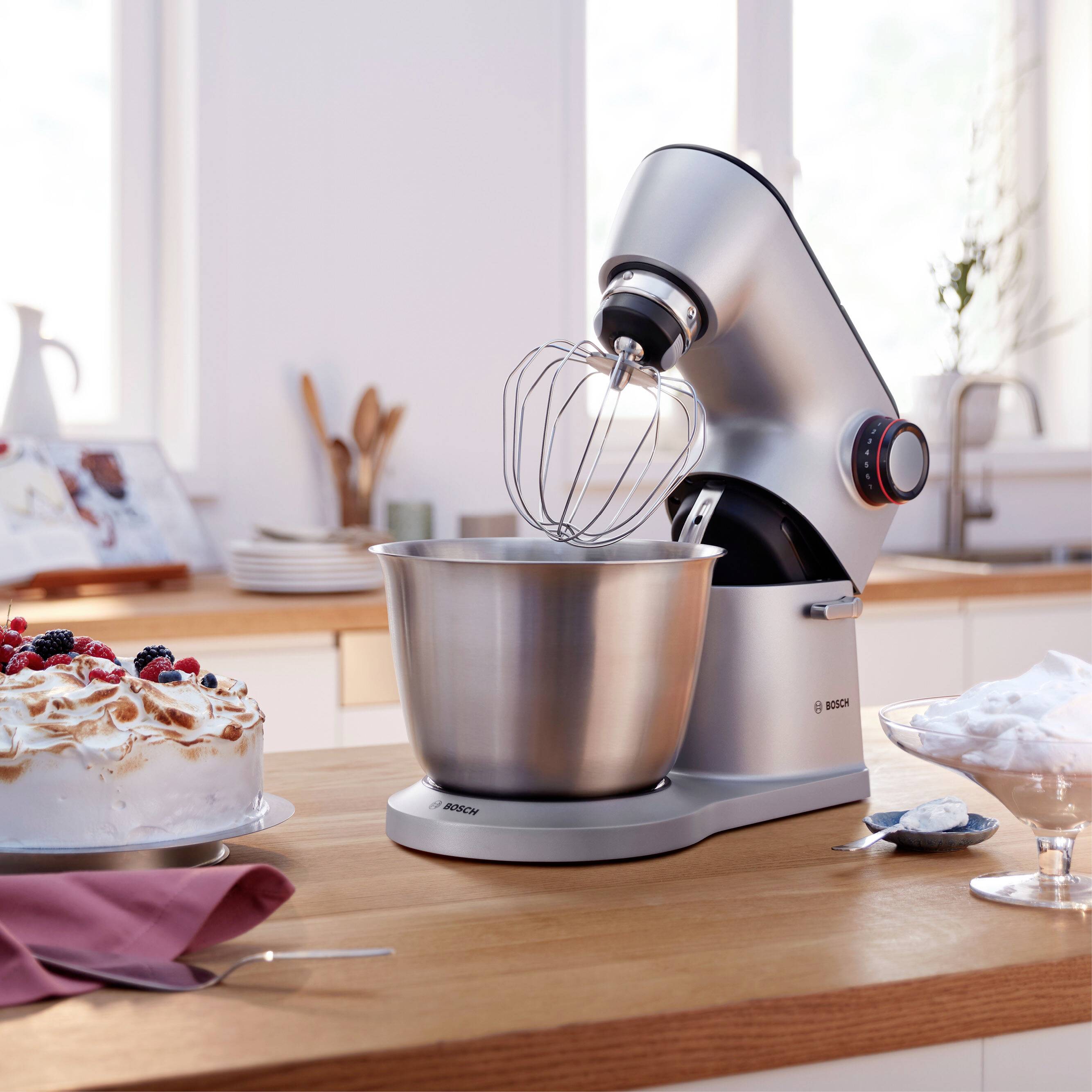 Srebrni kuhinjski robot na leseni mizi, pripravljen za mešanje. Ob njem smetanova torta z jagodami in steklena posoda z desertom.
