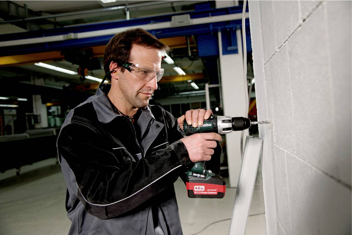 Moški v delovni obleki z akumulatorskim vijačnikom vrta luknjo v betonsko steno v delavniškem okolju.
