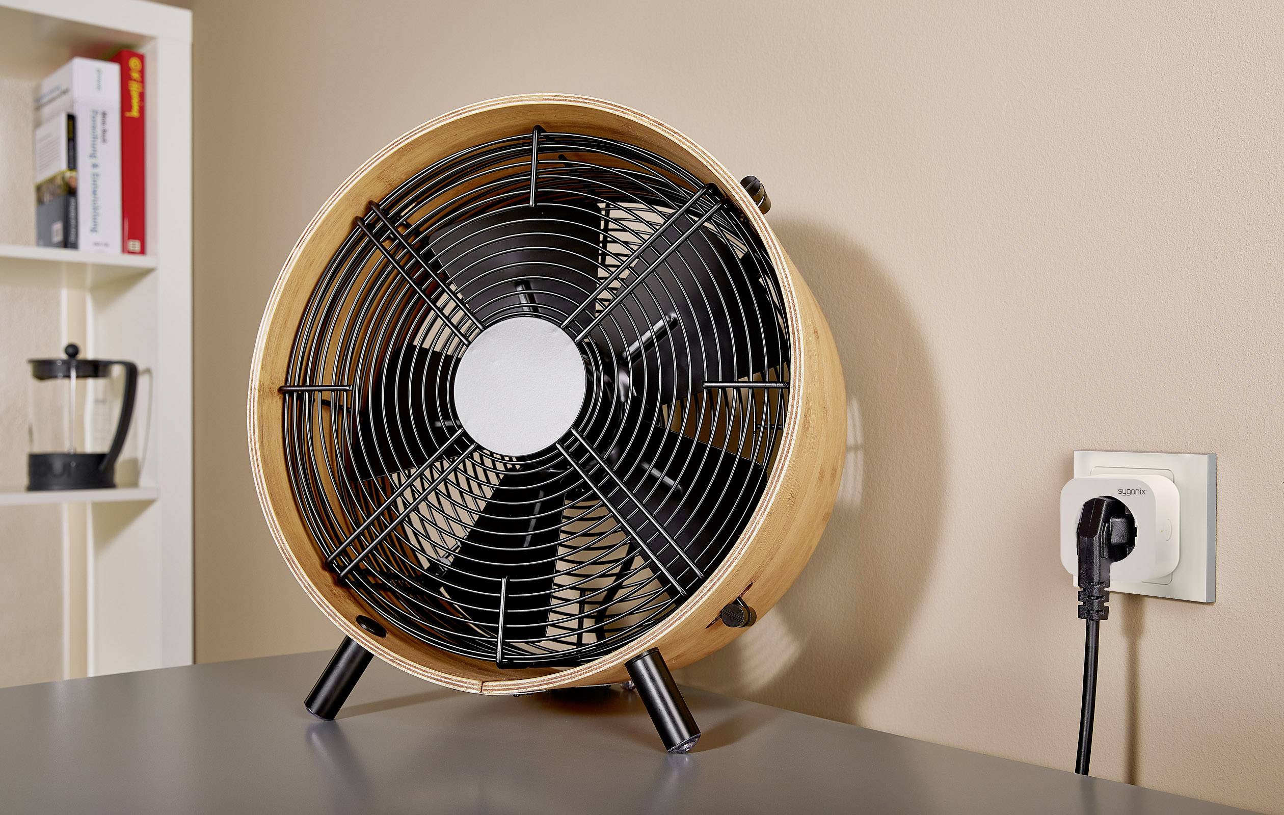 Okrogel namizni ventilator v videzu lesa stoji na mizi. Desno poleg njega je vtičnica z vtaknjenim vtičem. V ozadju je mogoče videti knjižno polico.