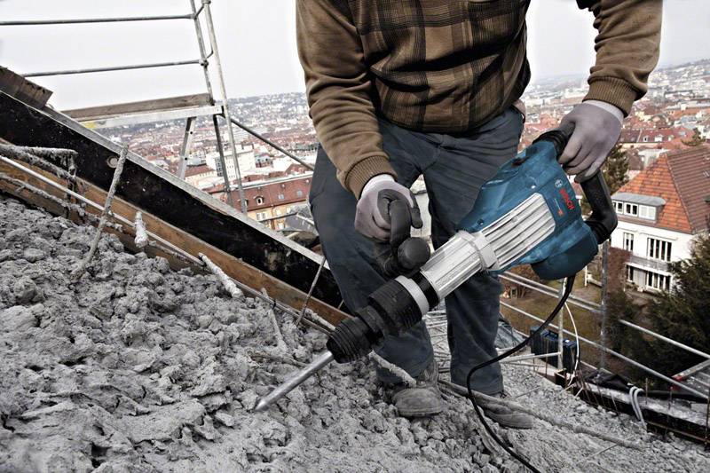 Oseba v delovni opremi uporablja pnevmatsko kladivo za rušenje betona na gradbišču, v ozadju pa se nahaja mesto.