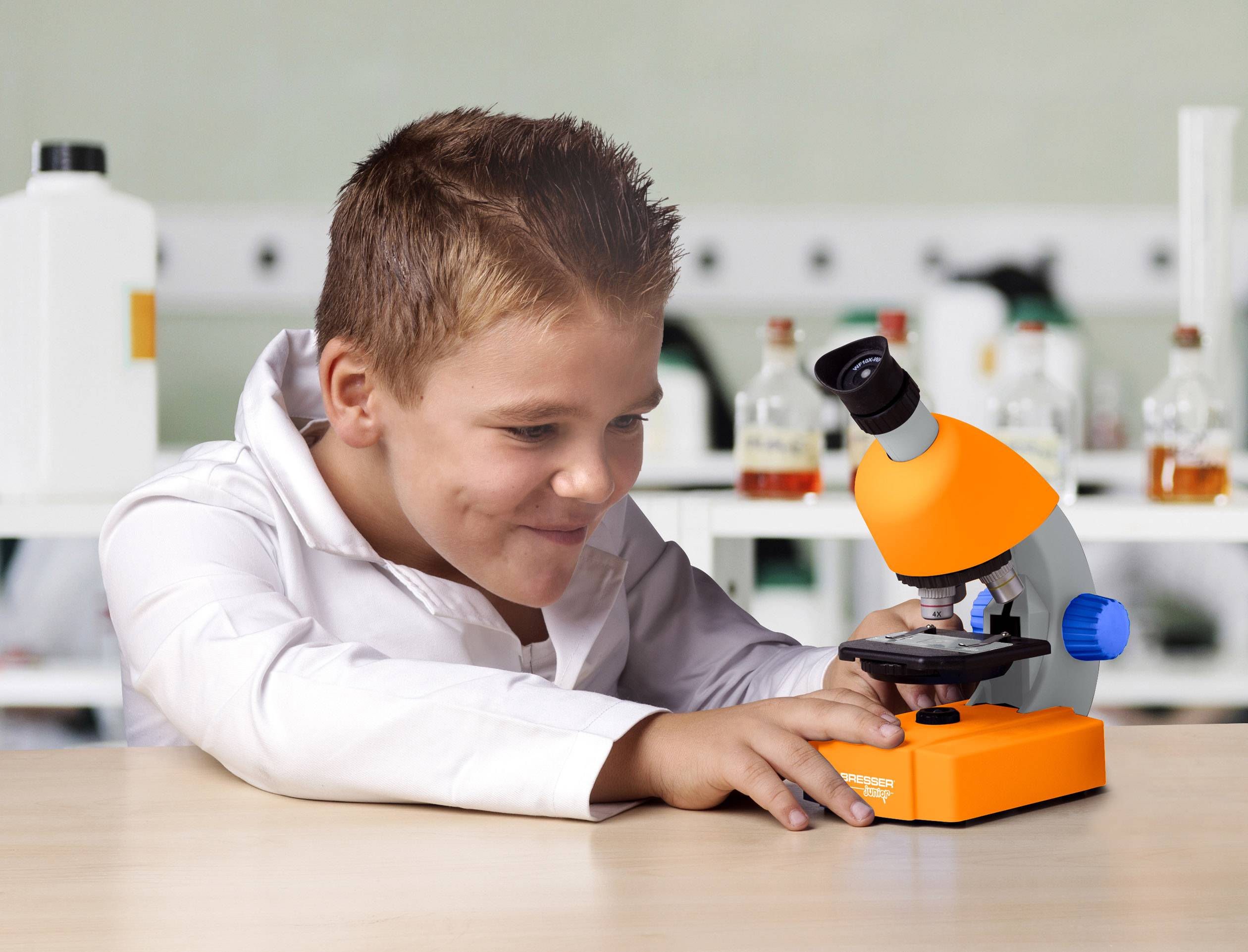 Deček opazuje predmet pod mikroskopom v laboratoriju. V ozadju so steklenice in laboratorijska oprema.
