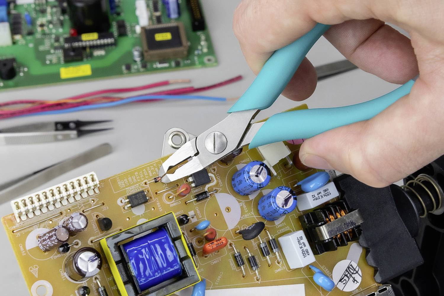 Roka drži klešče in popravlja elektronsko ploščo. V ozadju so vidna še druga orodja in plošče.