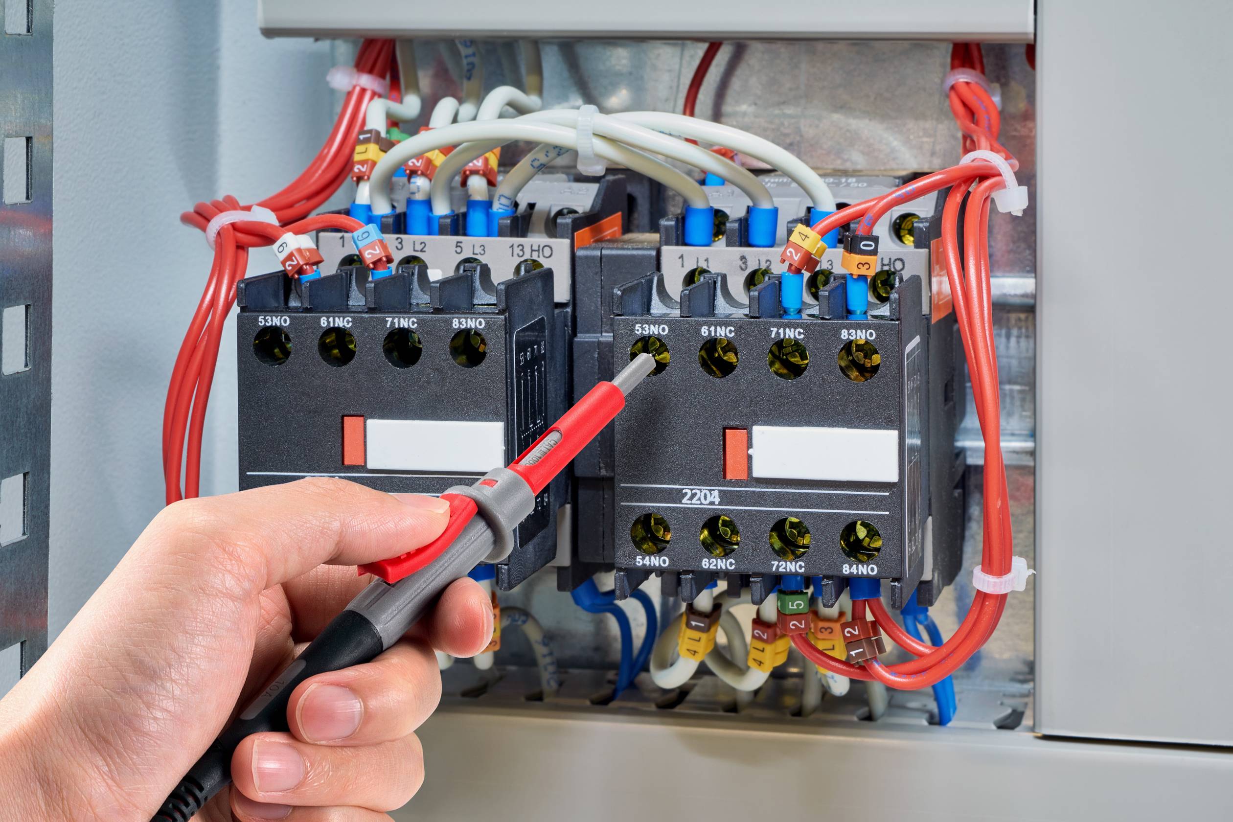 Roka drži napetostni preizkuševalnik v elektro omari. Različni pisani kabli so povezani z stikalno napravo.