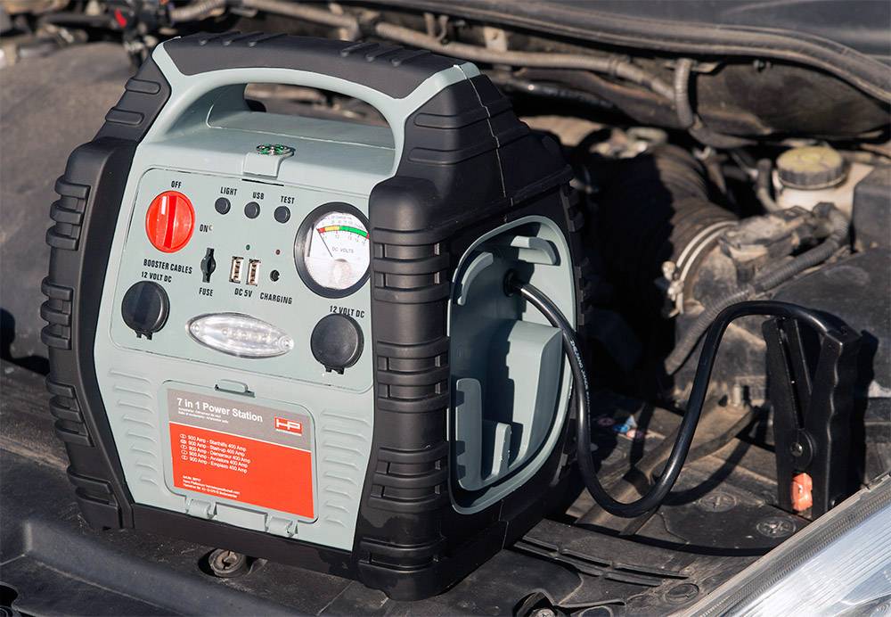 Prenosna startna naprava in powerbank z indikatorjem polnjenja in kabli, postavljena pred odprtim motornim prostorom, pripravljena za uporabo.