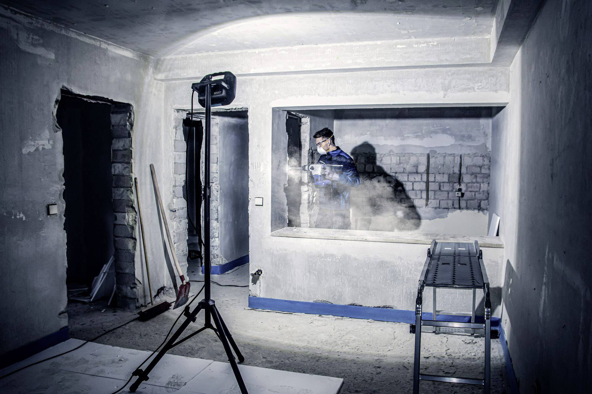 Oseba dela v nedokončanem prostoru na gradbišču. Gradbeni reflektor osvetljuje sceno. V ozadju so neometani zidovi.