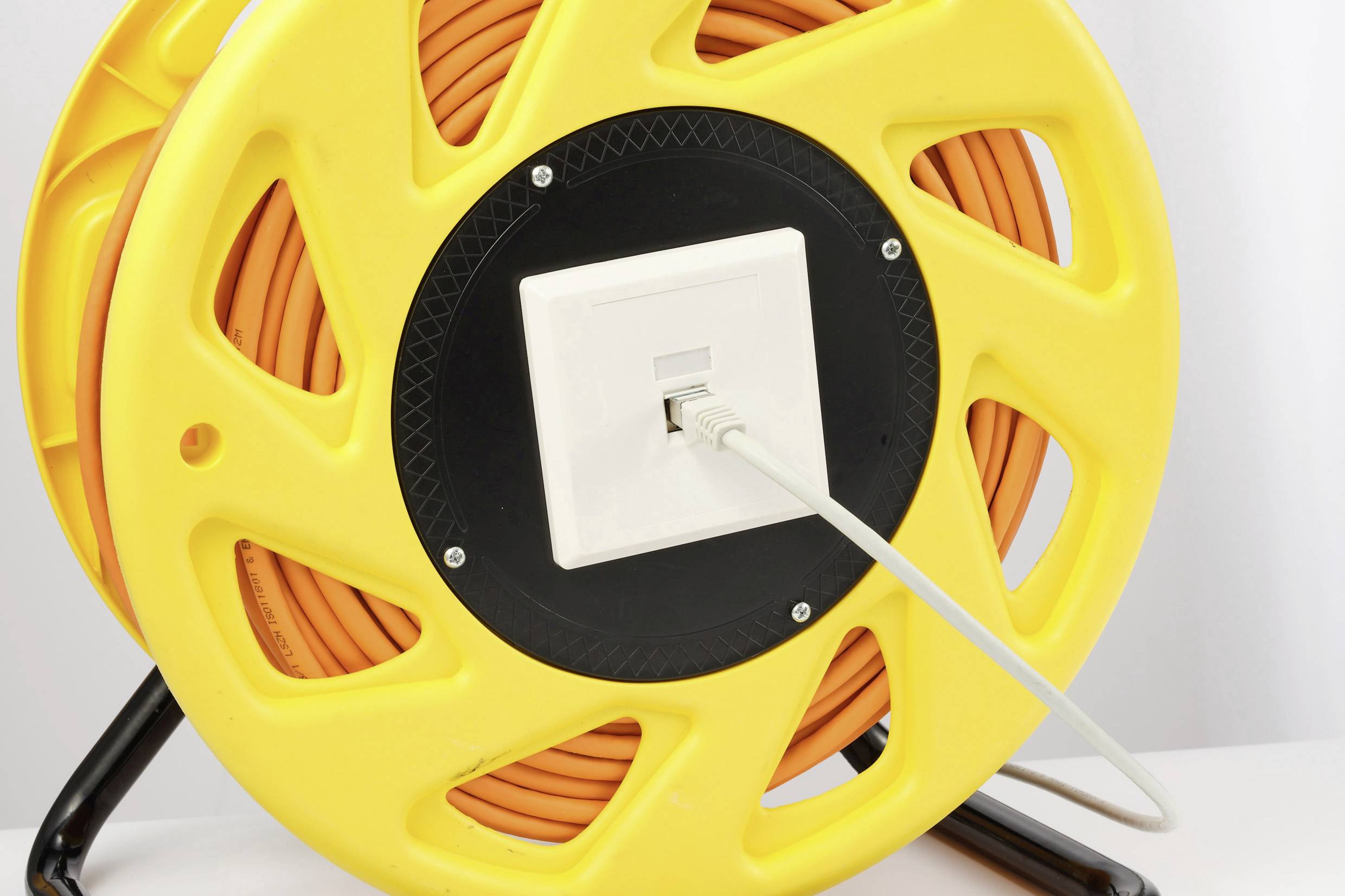Rumena kabelska kolut z navijanim oranžnim kablom, priključen je bel omrežni kabel. Stoji na kovinskem stojalu.