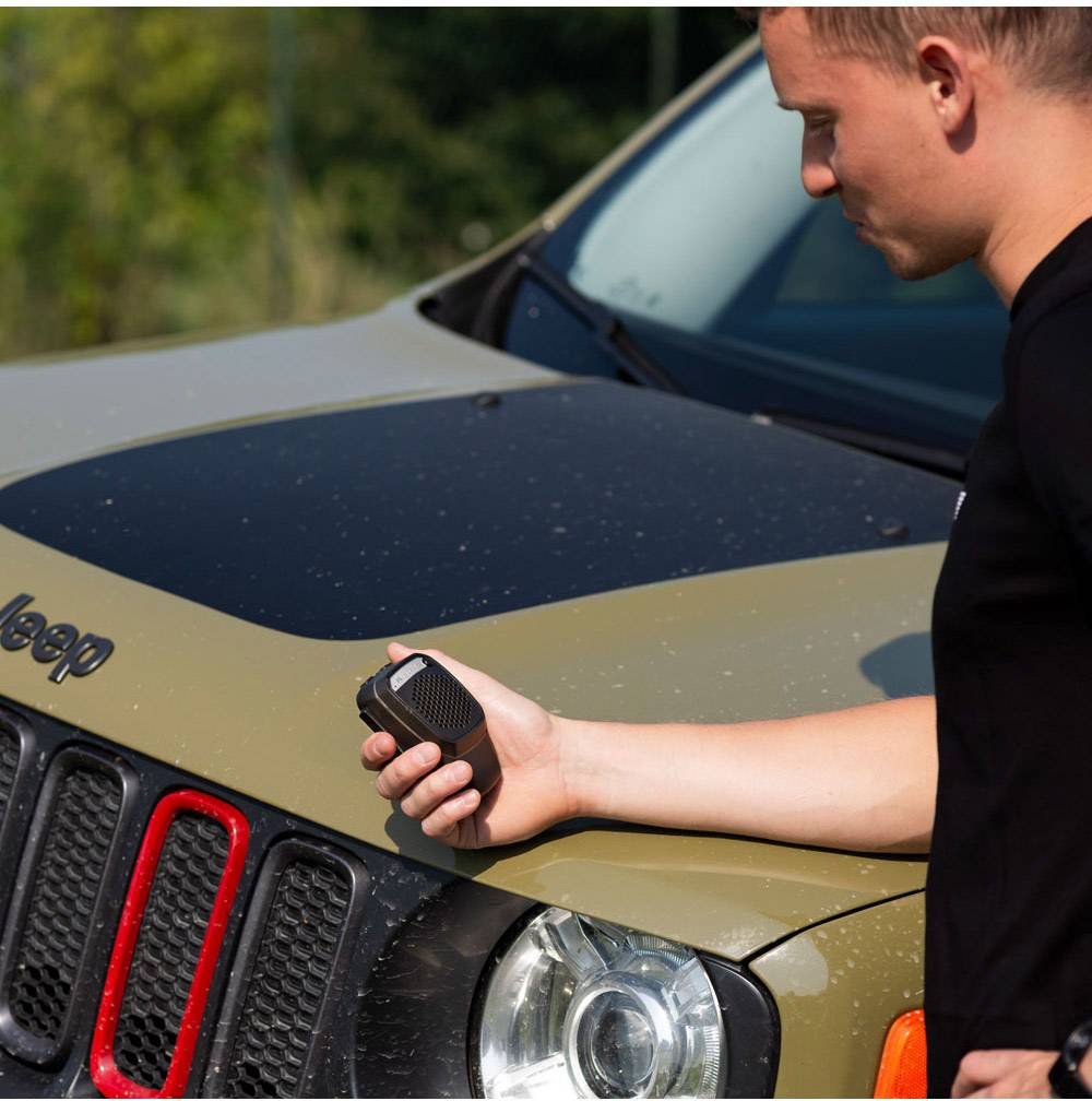 Moški stoji poleg Jeep-a in drži walkie-talkie. Jeep ima zeleno pokrov motorja in rdeče detajle na hladilniku.