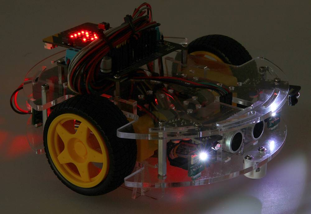 Majhen, prozoren robot na kolesih z rumenimi gumami, povezanimi električnimi vezji in svetlečimi LED-lučkami.