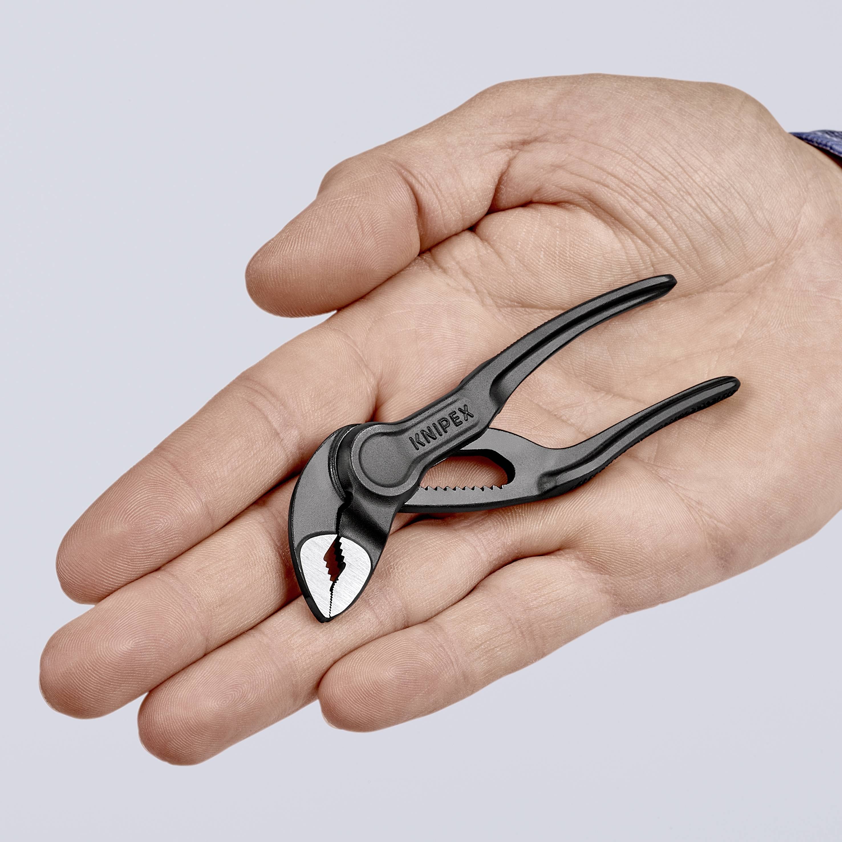 Ena roka drži majhne črne klešče z napisom 'KNIPEX'. Klešče so odprte in vidni so njihovi zobje.