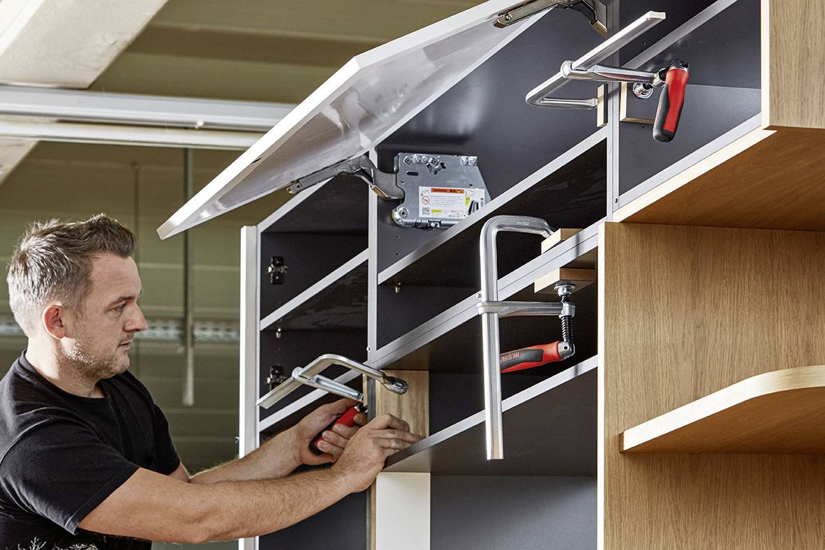 Moški z orodjem montira garderobne omar, pritjuje deske, da zagotovi funkcionalnost, pri delu pa je zelo skrben.
