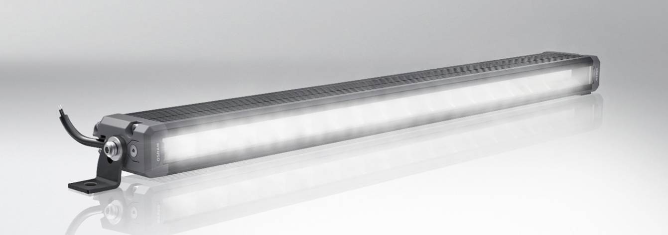 Podolgovata LED-svetilka s črnim ohišjem in kabelskim priključkom, nameščena na nosilcu. Nežno osvetljuje površino.