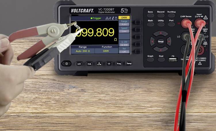 Digitalni multimeter na leseni mizi prikazuje '999.809'. Merilni kabli s sponkami so priključeni. Levo je roka med merjenjem.