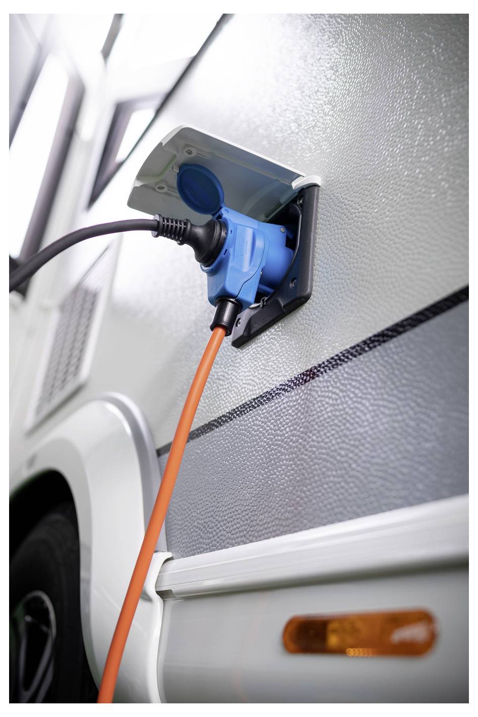 Detaljni pogled na prikolico ali kamp prikolico, ki je priključena na električno vtičnico s porta pomarančnim napajalnim kablom, povezanim z modrim adapterjem za vtič.