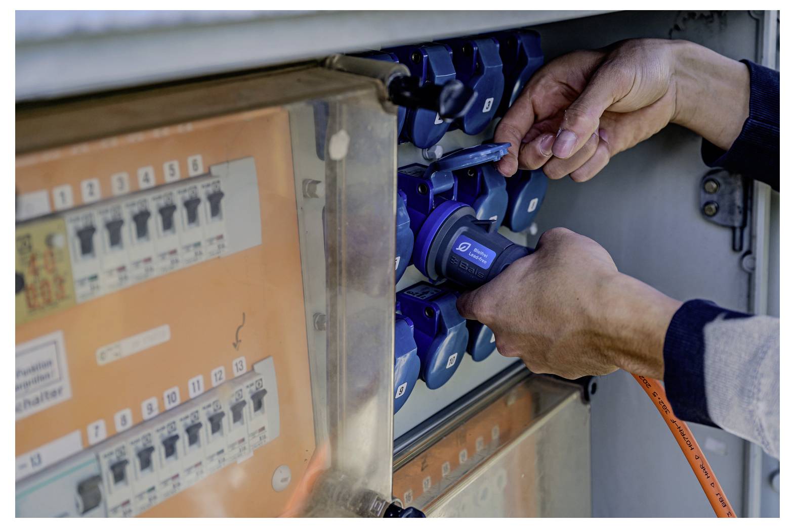 Roke, ki vstavljajo oranžni napajalni kabel v modri industrijski električni priključek na elektroenergetski plošči.