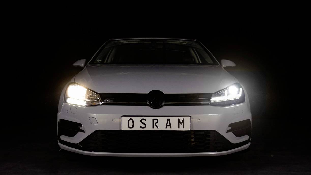 Belo vozilo z črno masko hladilnika in prižganimi LED-žarometi stoji v temnem prostoru. Registrska tablica prikazuje 'OSRAM'.
