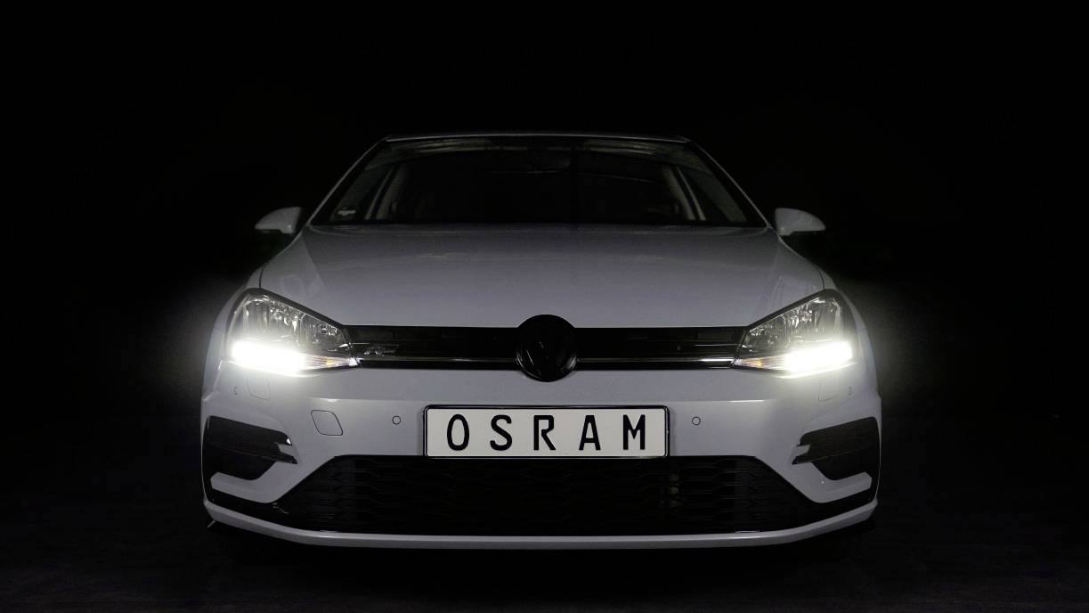 Avto z prižganimi žarometi na temnem ozadju. Registrska tablica z napisom 'OSRAM'.
