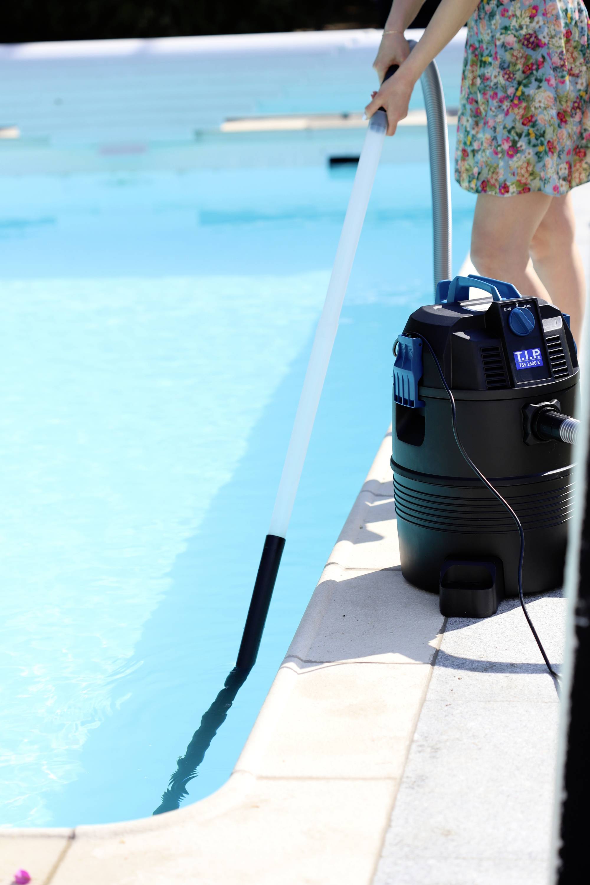 Oseba čisti rob bazena s čistilcem bazena. Sesalec stoji ob robu bazena in je povezan s kablom.