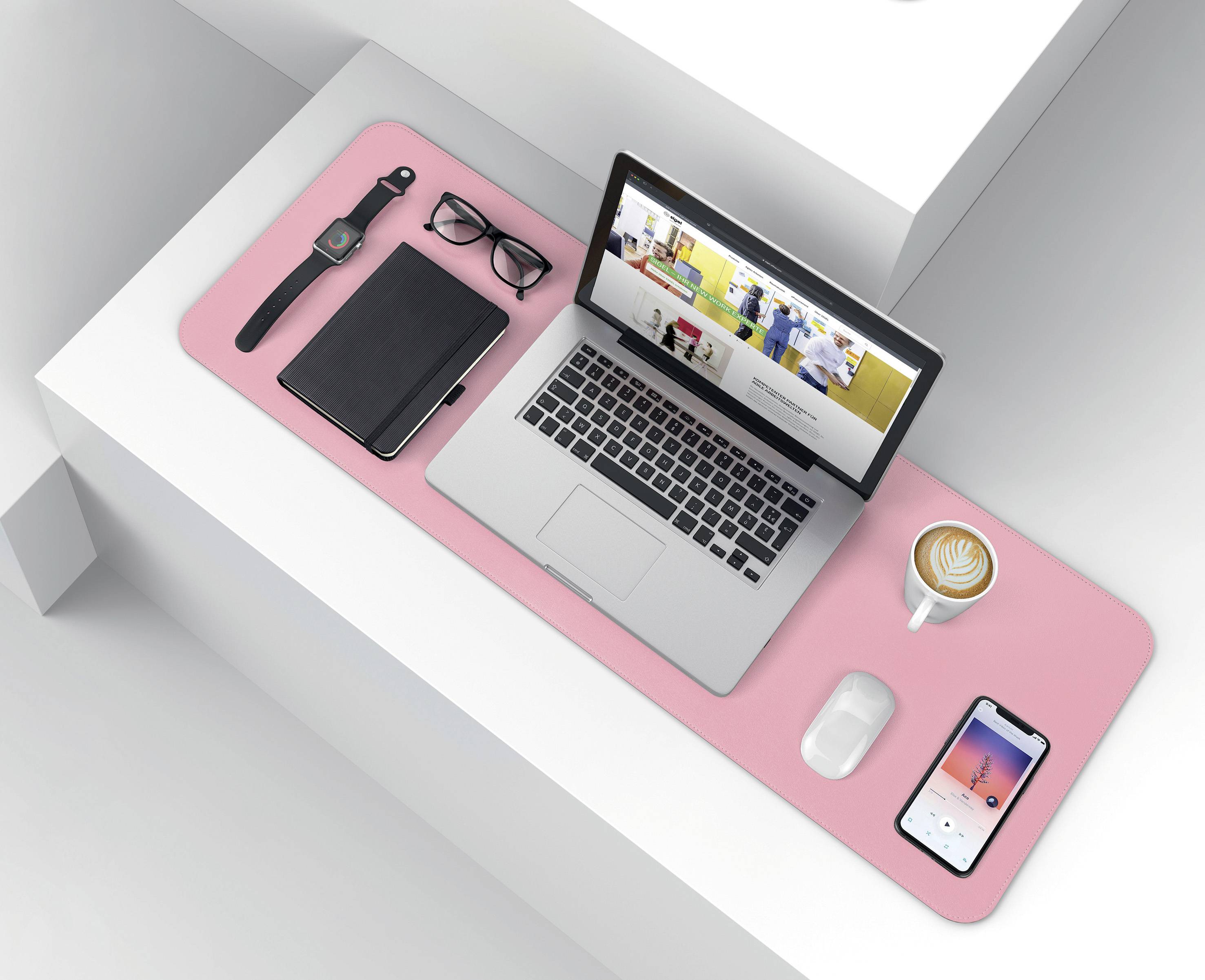 Prenosnik na rožnati podlogi za pisalno mizo, zraven beležka, očala, pametna ura in pametni telefon. Skodelica kave ob strani, na zaslonu odprta spletna stran.