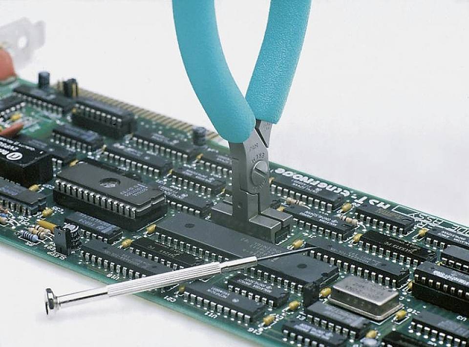 Klešče obdelujejo tiskano vezje z veliko elektronskimi komponentami, zraven leži majhen izvijač.