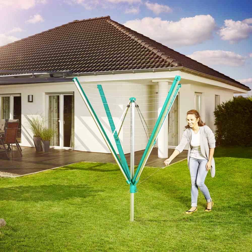 Ženska obešuje perilo na zelen sušilni stativ v sončnem vrtu, v ozadju moderna hiša.