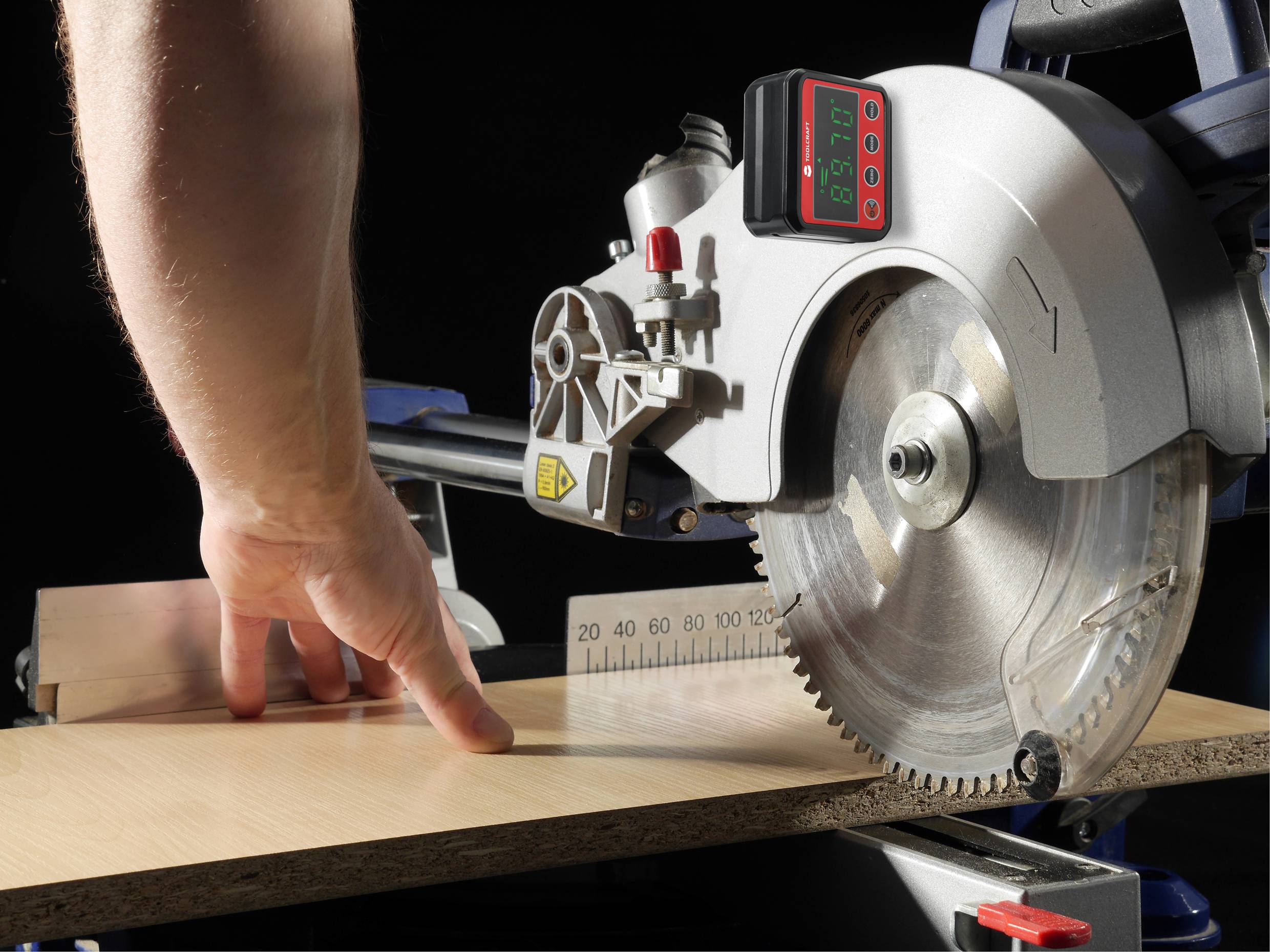 Ena roka drži leseno desko pred krožno žago, pripravljena za rezanje. Digitalna merilna naprava prikazuje kot.